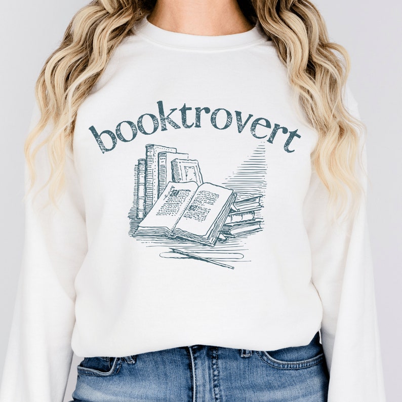 white unisex booktrovert shirt that says booktrovert in navy blue writing along with a graphic of books, perfect bookish merch for book lovers