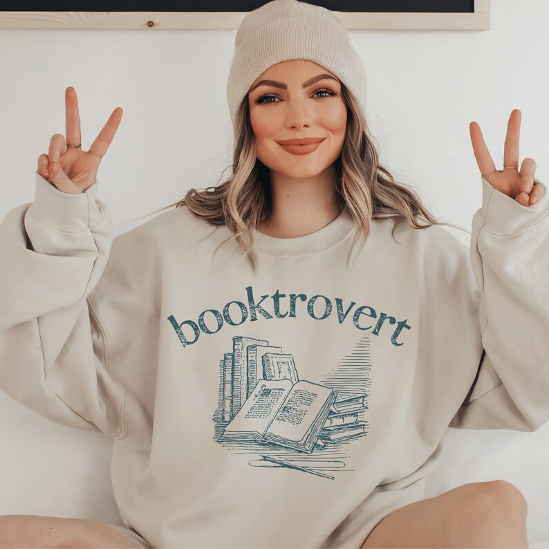 beige unisex booktrovert shirt that says booktrovert in navy blue writing along with a graphic of books, perfect bookish merch for book lovers