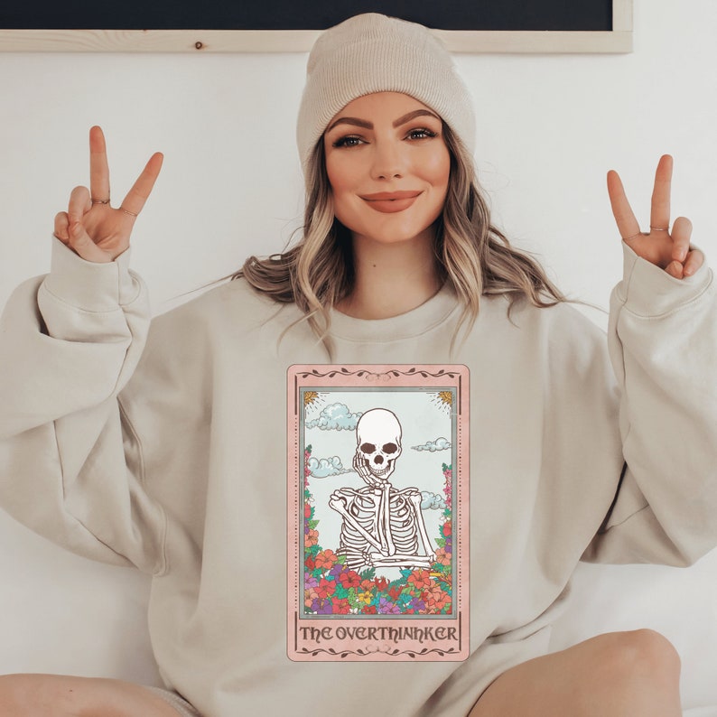 beige unisex overthinker sweatshirt with a graphic of a tarot card with skeleton graphic that says the overthinker
