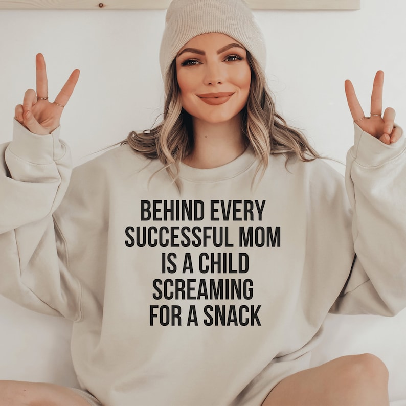 white unisex funny mom sweatshirt on female model that says behind every successful mom is a child screaming for a snack in black lettering. Perfect gift for a mompreneur or working mom.