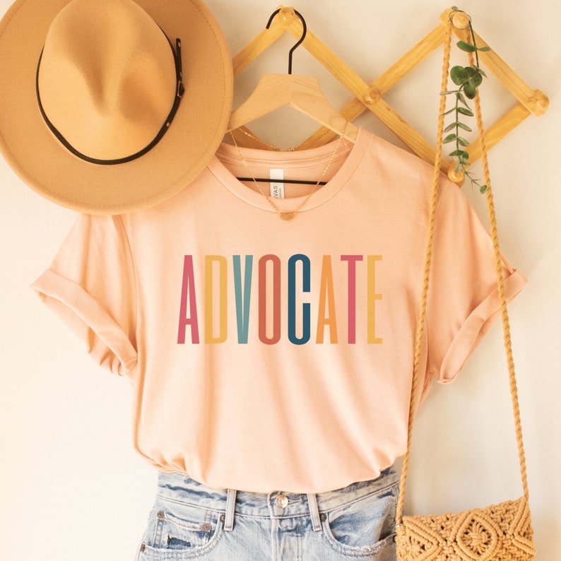 heather peach unisex t-shirt that says the word "advocate" in capital, multicolored letters