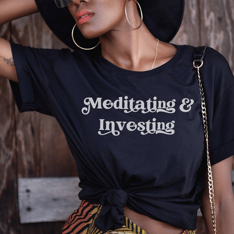 Black unisex t-shirt on female model with a design that says "meditating and investing" in white lettering with a unique font. Perfect gift for someone interested in personal finance, meditation, and investing.  