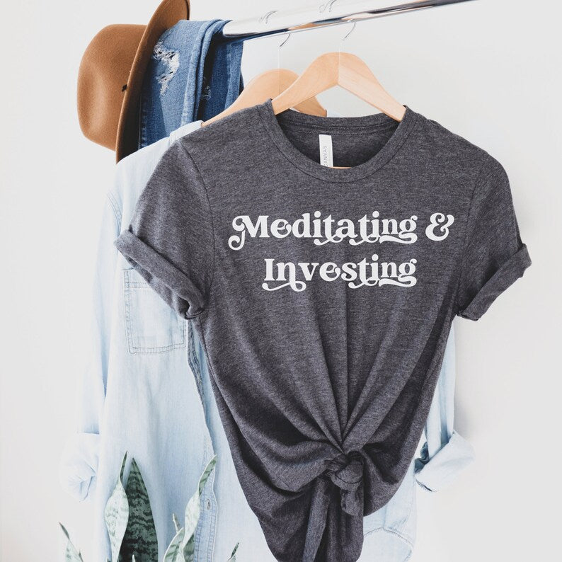 dark heather gray unisex t-shirt on female model with a design that says "meditating and investing" in white lettering with a unique font. Perfect gift for someone interested in personal finance, meditation, and investing. 