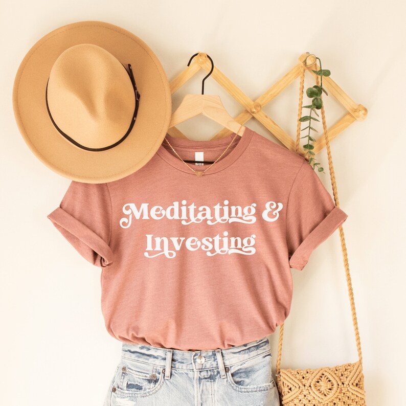 heather mauve unisex t-shirt on female model with a design that says "meditating and investing" in white lettering with a unique font. Perfect gift for someone interested in personal finance, meditation, and investing. 