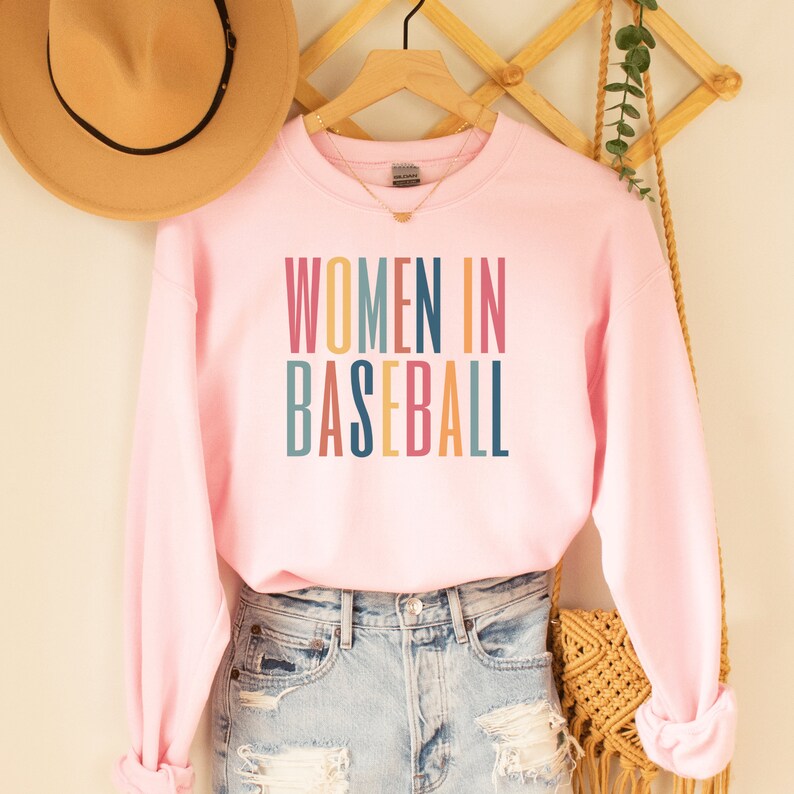 Pink unisex sweatshirt with multicolored letters that say women in baseball in all caps