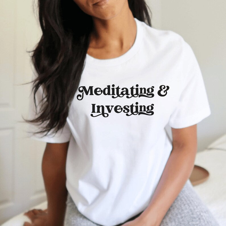 white unisex t-shirt on female model with a design that says "meditating and investing" in black lettering with a unique font. Perfect gift for someone interested in personal finance, meditation, and investing. 