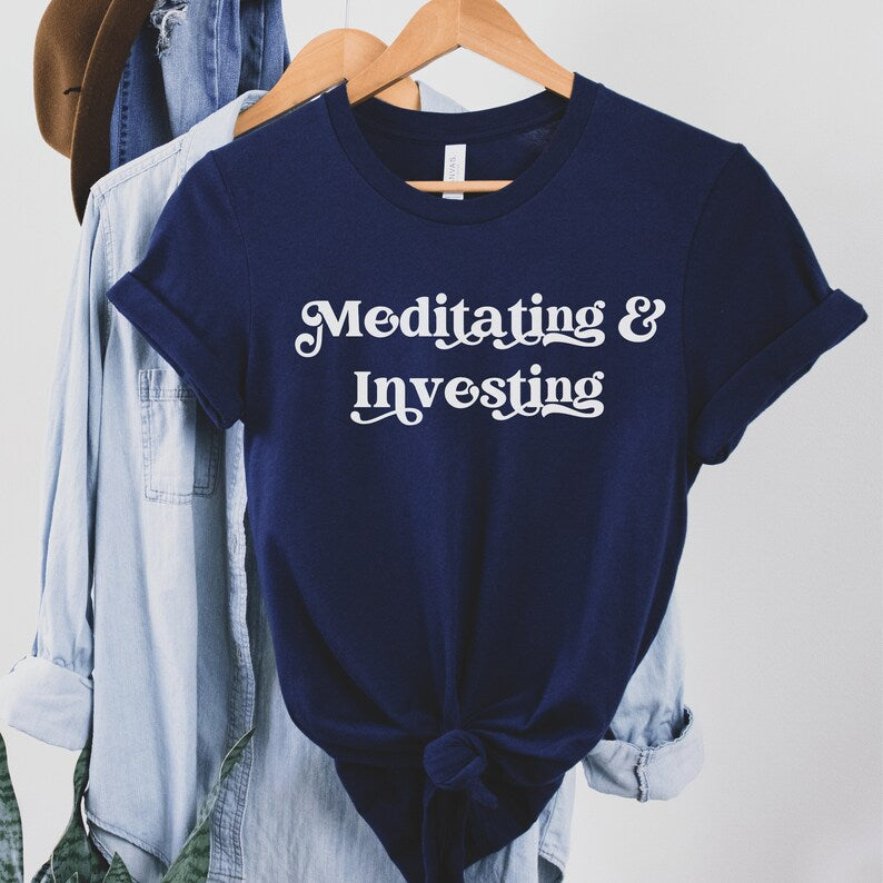 Navy blue unisex t-shirt on female model with a design that says "meditating and investing" in white lettering with a unique font. Perfect gift for someone interested in personal finance, meditation, and investing. 