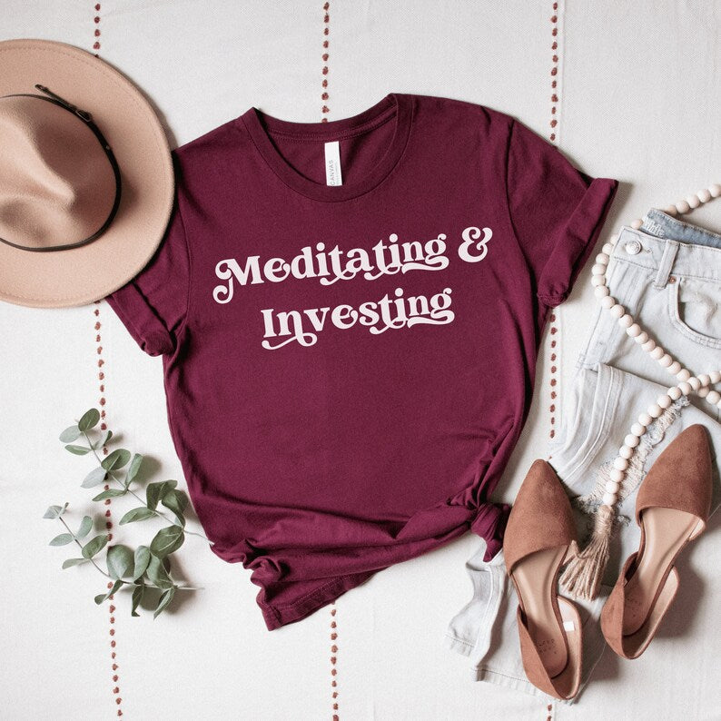 Maroon unisex t-shirt on female model with a design that says "meditating and investing" in white lettering with a unique font. Perfect gift for someone interested in personal finance, meditation, and investing. 