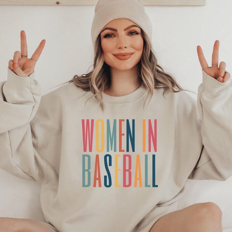 Beige unisex sweatshirt with multicolored letters that say women in baseball in all caps