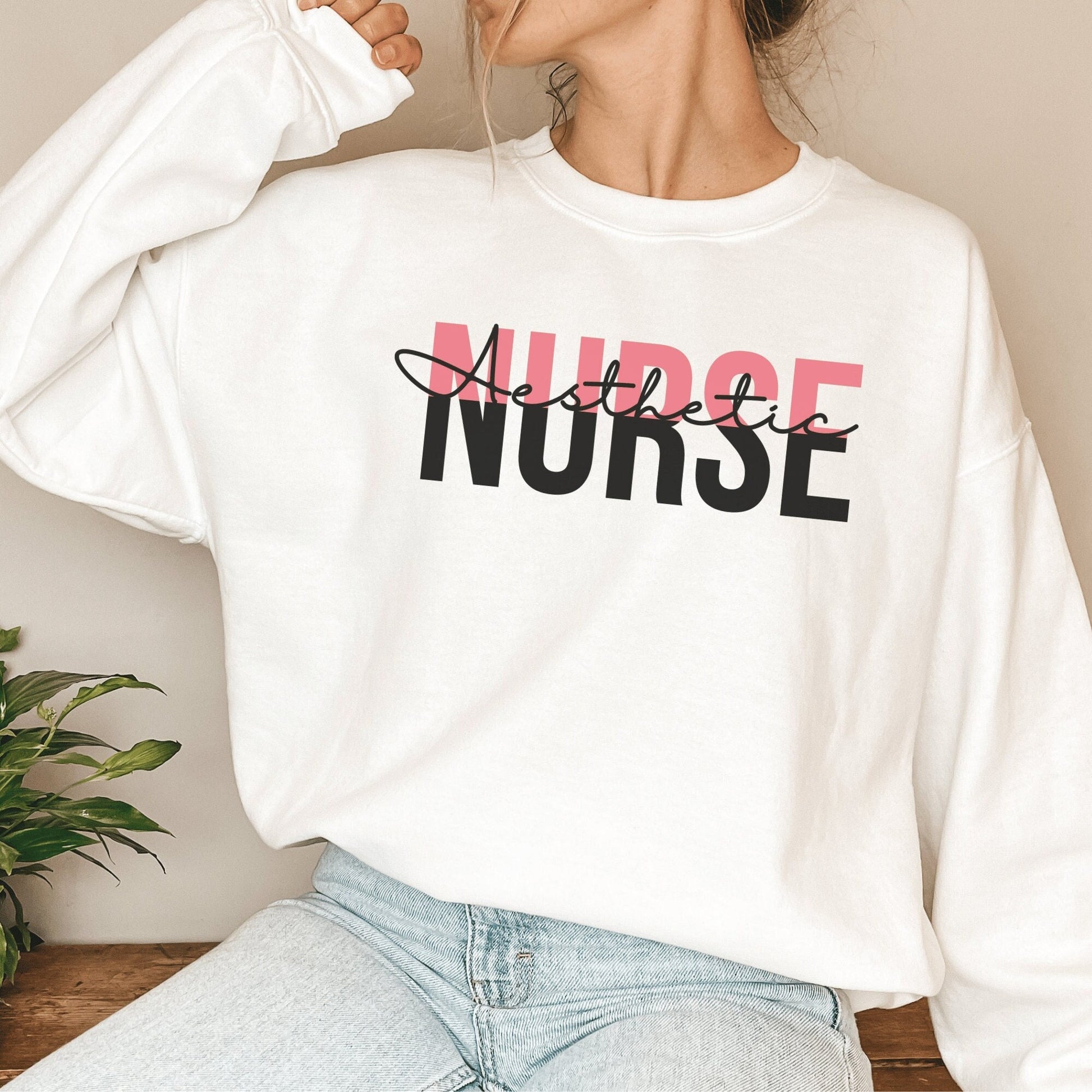 Aesthetic Nurse is a white unisex sweatshirt on a female model with the design in pink and black lettering that says Aesthetic Nurse. Aesthetic is in black cursive lettering and layered on top of the word nurse, which is in capital letters that are pink and black.
